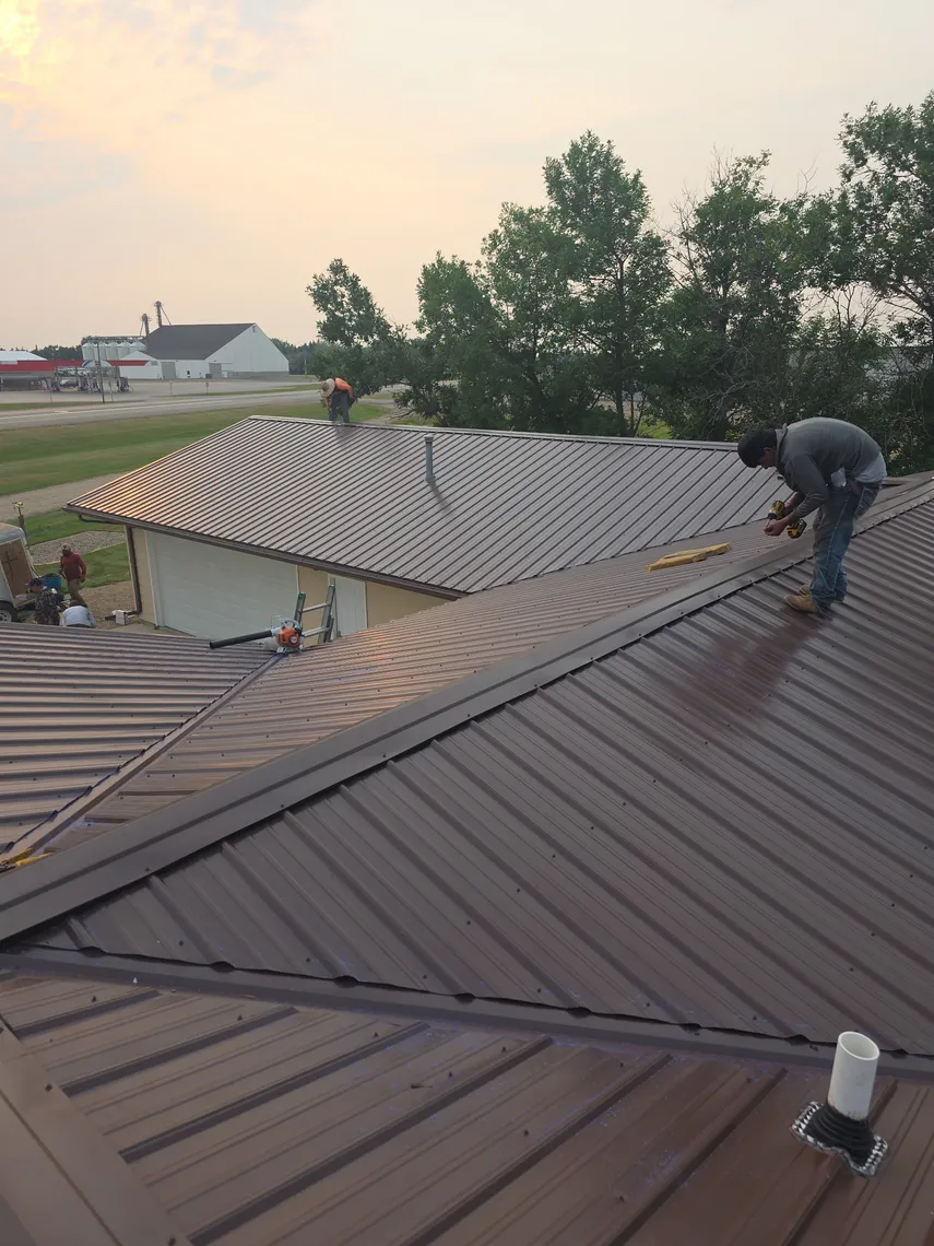 Aerial view of a Tops Roofing crew installing a brown standing-seam metal roof on a house near a grain elevator