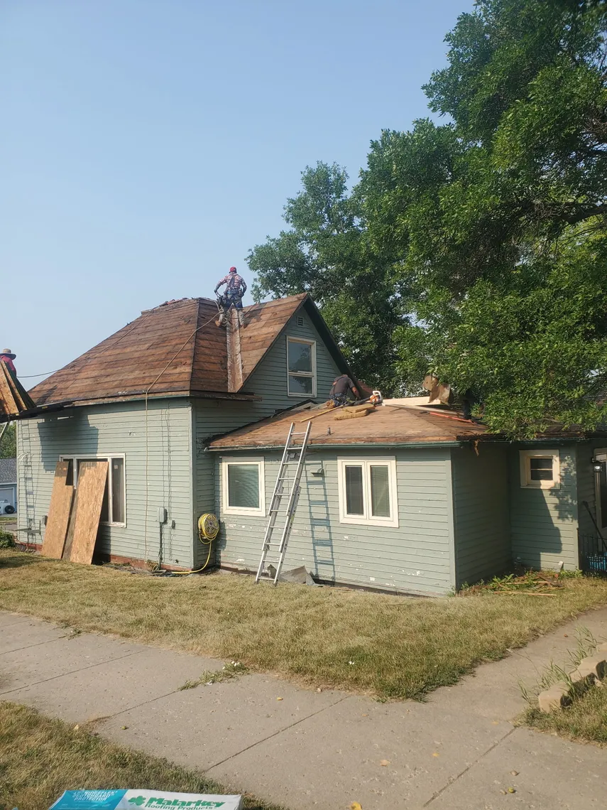 A Tops Roofing crew member tearing off old shingles down to the roof deck on a light green craftsman-style home