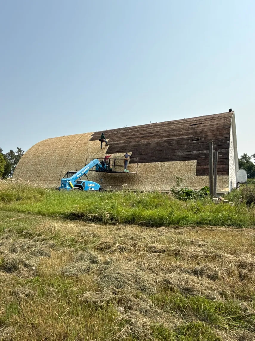A Tops Roofing crew on a boom lift installing new plywood decking over the curved roof of a Quonset barn