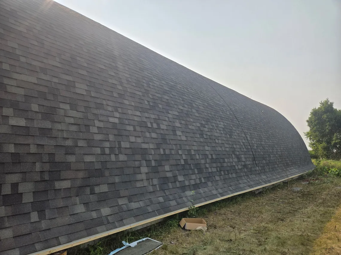 A finished gray asphalt-shingle roof sweeping over the curved arch of a Quonset-style barn
