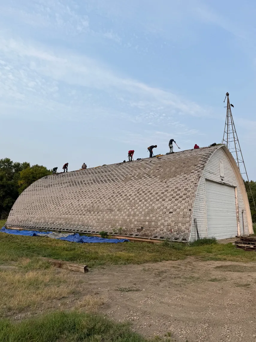 A crew tearing off the old shingle roof of a curved Quonset-style barn, working along its arched ridge