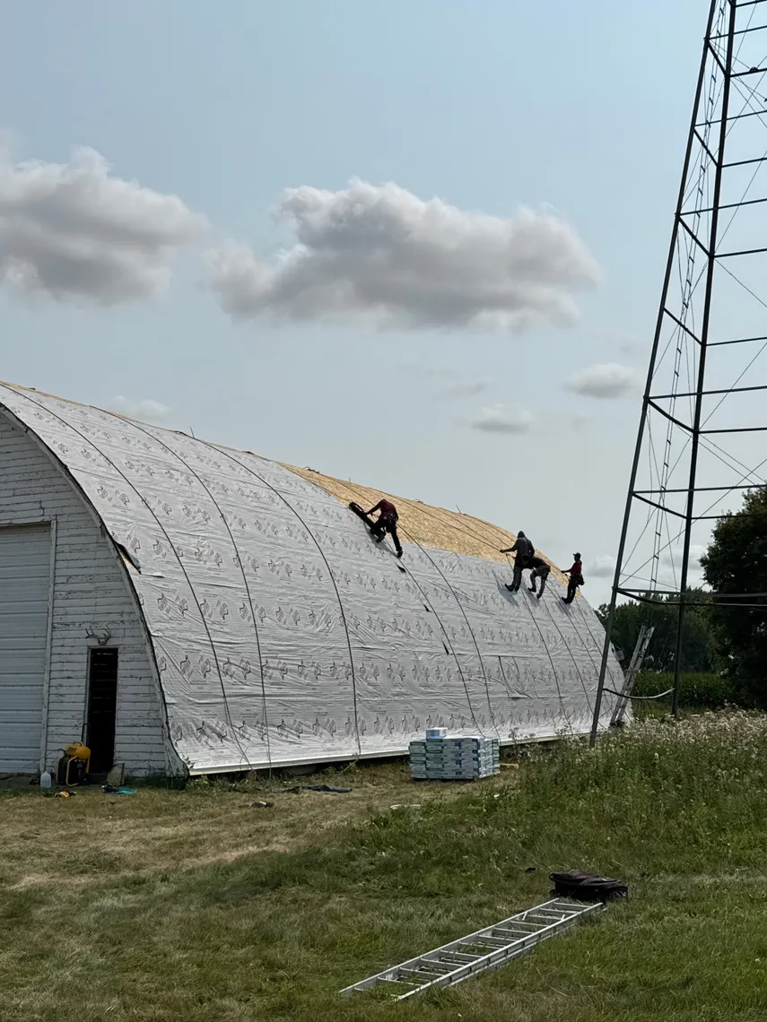 Crew members installing synthetic underlayment and starting new shingles on the arched roof of a Quonset barn