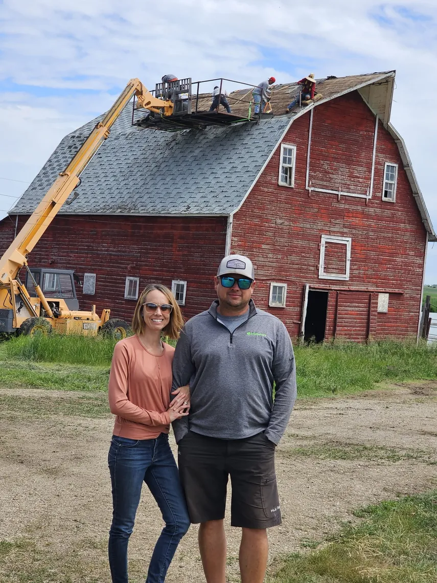 Two people in front of a red barn as a Tops Roofing crew tears off its old shingle roof from a boom lift platform